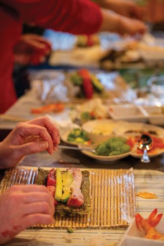 Hands hover above an unrolled piece of sushi, with ingredients like fish and avocado visible