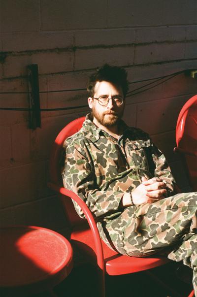 Color photo of Rock Eupora main man Clayton Waller, wearing a camo jumpsuit and sitting outside on a red metal patio chair against a cinder block wall.