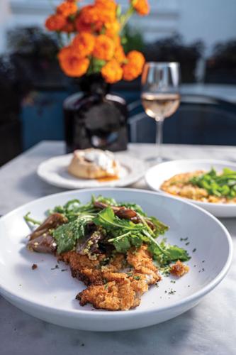 A breaded veal cutlet under leafy greens, plated near a glass of wine and orange flowers