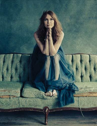 Color photo of the artist facing the camera and looking off to her left, wearing a sheer green-blue dress and resting her chin on her hands with her arms propped on her knees, sitting atop the back of a green couch against a gray background.