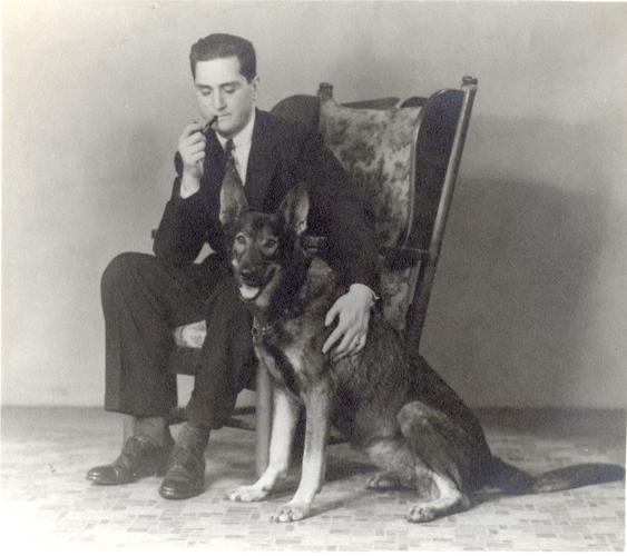 A black-and-white photo of a man in an armchair, smoking and pipe and petting a German shepherd