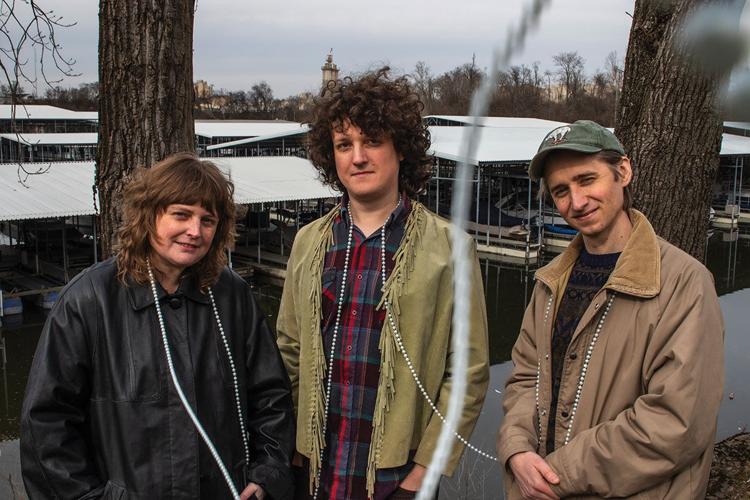 Color press photo of the three band members standing outside, wearing winter coats, with a marina in the background.