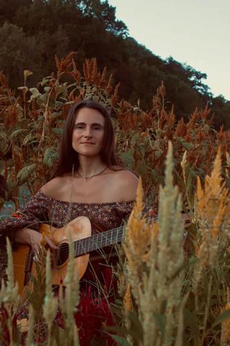 Color press photograph of musician Jessie Gray, wearing a dark red patterned off-the-shoulder dress and turquoise jewelry, playing a nylon-string guitar amid some sagebrush.