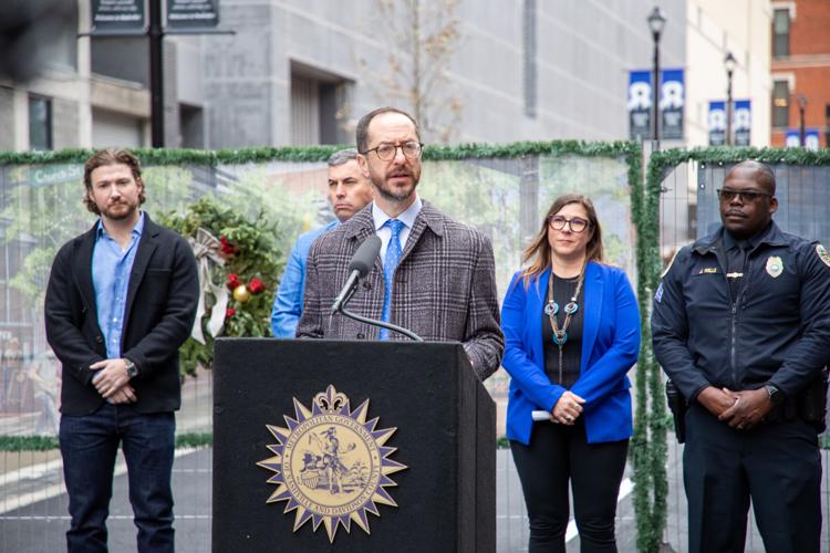Freddie O'Connell stands at a podium on a city street, four people including a uniformed police officer stand behind them, a barricade fencing off the street