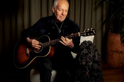 Color photo of the artist, dressed in black, playing an acoustic guitar as a labradoodle looks on