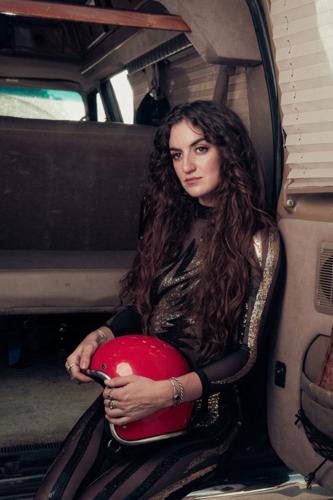 Color press photo of singer-songwriter Chloe Kimes for her ablum 'Daredevil,' dressed in a catsuit-type stage costume, holding a motorcycle helmet and sitting in the back of a van,
