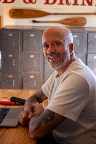 A man in a white t-shirt sits in front of an open laptop, arms crossed, with a set of lockers behind him and a pair of crossed oars above them.