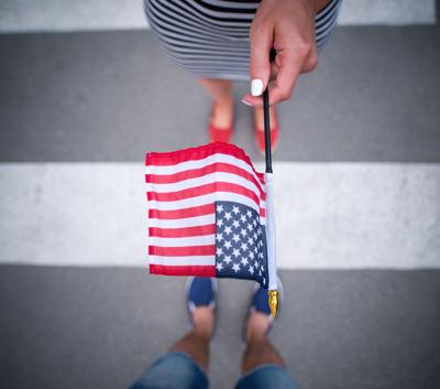 A stock image of a person holding a small American flag, shot with the camera pointing down to a line on the pavement that the person is standing behind.