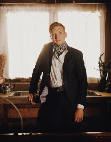 Clad in a dark suit jacket and a black and white neckerchief, artist stands by a kitchen sink in front of a window and looks through the camera back into the room