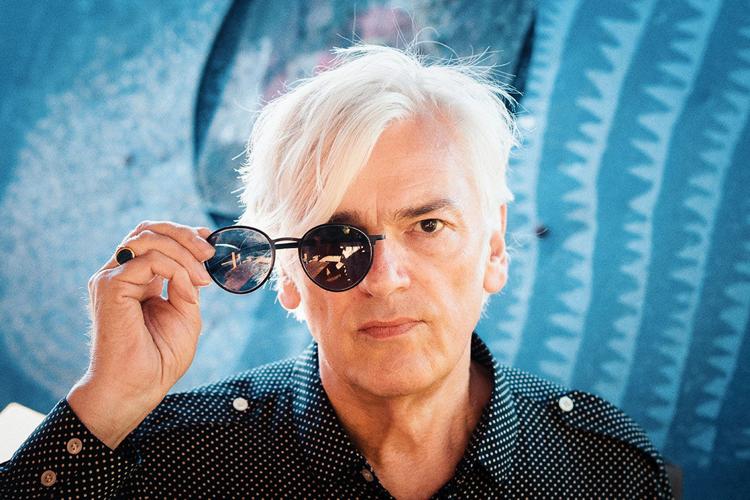 Color press photo of Robyn Hitchcock looking sternly through one eyepiece of a pair of sunglasses. The effect is as if he's stepped part way out from behind the sunglasses as they hang in midair.