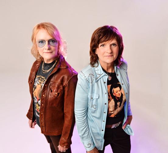 Color studio photo of Indigo Girls' Amy Ray and Emily Saliers, dressed in folk-rock stage wear of leather jackets and denim overshirts.