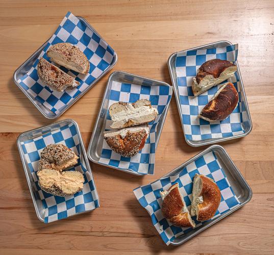 Color photo of several bagel-and-schmear combinations laid out on a table, each in their own tray with blue and white checked paper underneath