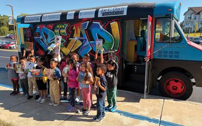 A crowd of children holds up picture books in front of a colorful bus with "Read" emblazoned on it.