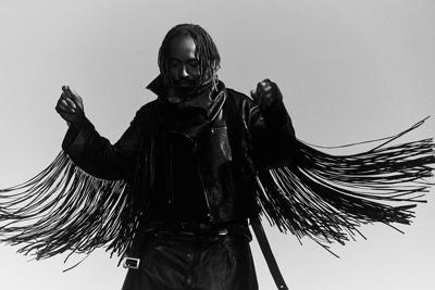 Black and white press photo of bass virtuoso Thundercat, wearing a jacket with very long fringe dangling from the arms, facing the camera while raising his arms and looking down