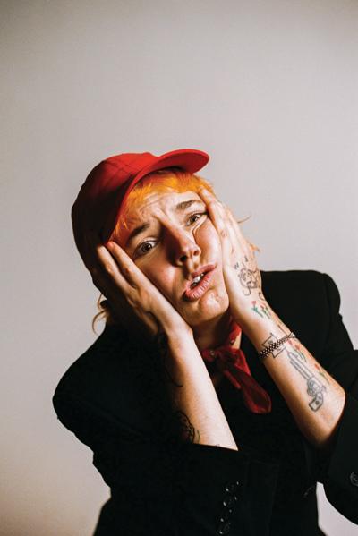 Wearing a black long-sleeve shirt and a red cap, the artist poses in front of a white background with their hands to the side of their face in an expression of dismay and exhaustion.