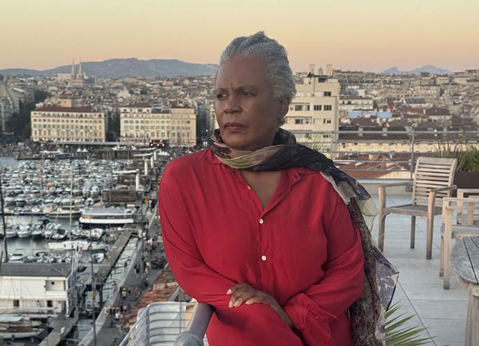 Author press photo of Claudia Rankine, wearing a red suit and a black silk scarf, looking out to the camera's left off a balcony. A busy harbor and the city beyond are visible behind her.