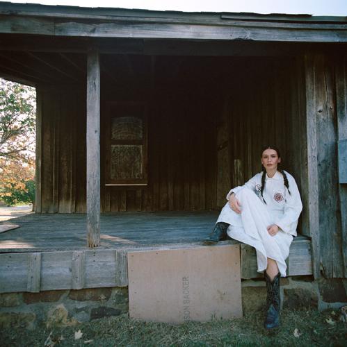 Wearing a long white dress and cowboy boots, artist sits on the porch of a wooden structure and looks into the camera.