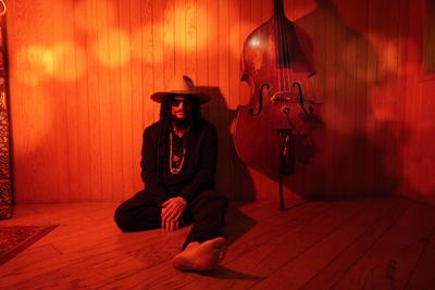 Barefoot, wearing a wide-brim hat and sunglasses and lit in a shade of deep red, Don Was sits on a wooden floor next to an upright bass
