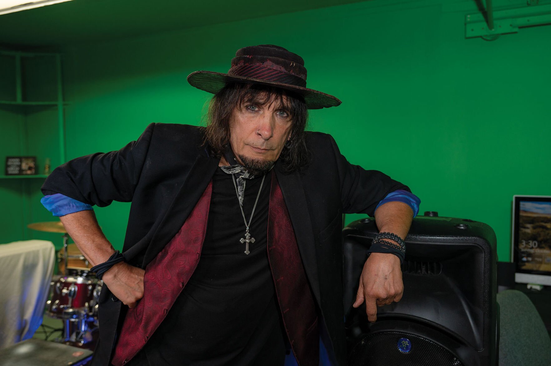 A man in. a brimmed hat and sports coat with the sleeves rolled rests his arm on a speaker in a green room.