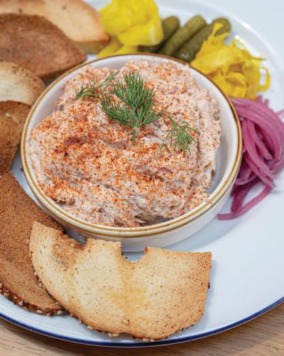 A small bowl of fish dip surrounded by pieces of toast and pickled vegetables.