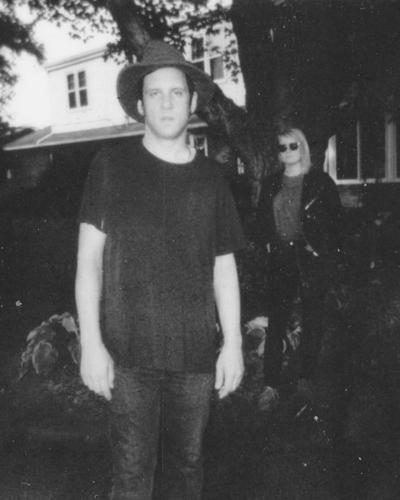 Black and white photo of singer-songwriter Ryan Davis standing near a tree outside a white house. A woman in sunglasses and a black leather jacket stands in the background.