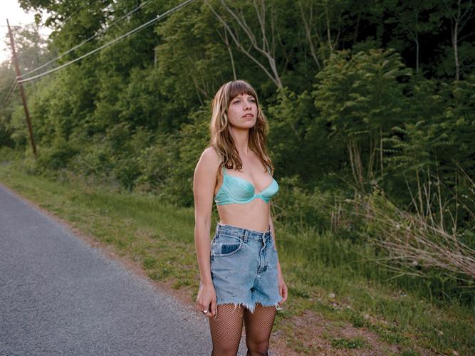 Color photo of the artist standing on a paved road near woods, looking at something off to the right of the image, wearing faded blue cut-off denim shorts, fishnet hose and a bright blue bra.