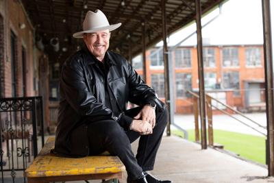 Wearing a black leather jacket and a white cowboy hat, Stacy Mitchhart sits on a wooden bench in a covered outdoor space and smiles into the camera.