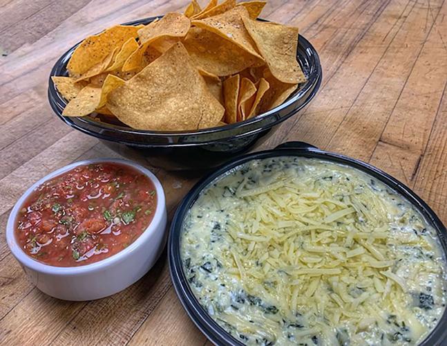 A bowl of artichoke dip topped with shredded cheese next to a basket of tortilla chips and bowl of salsa