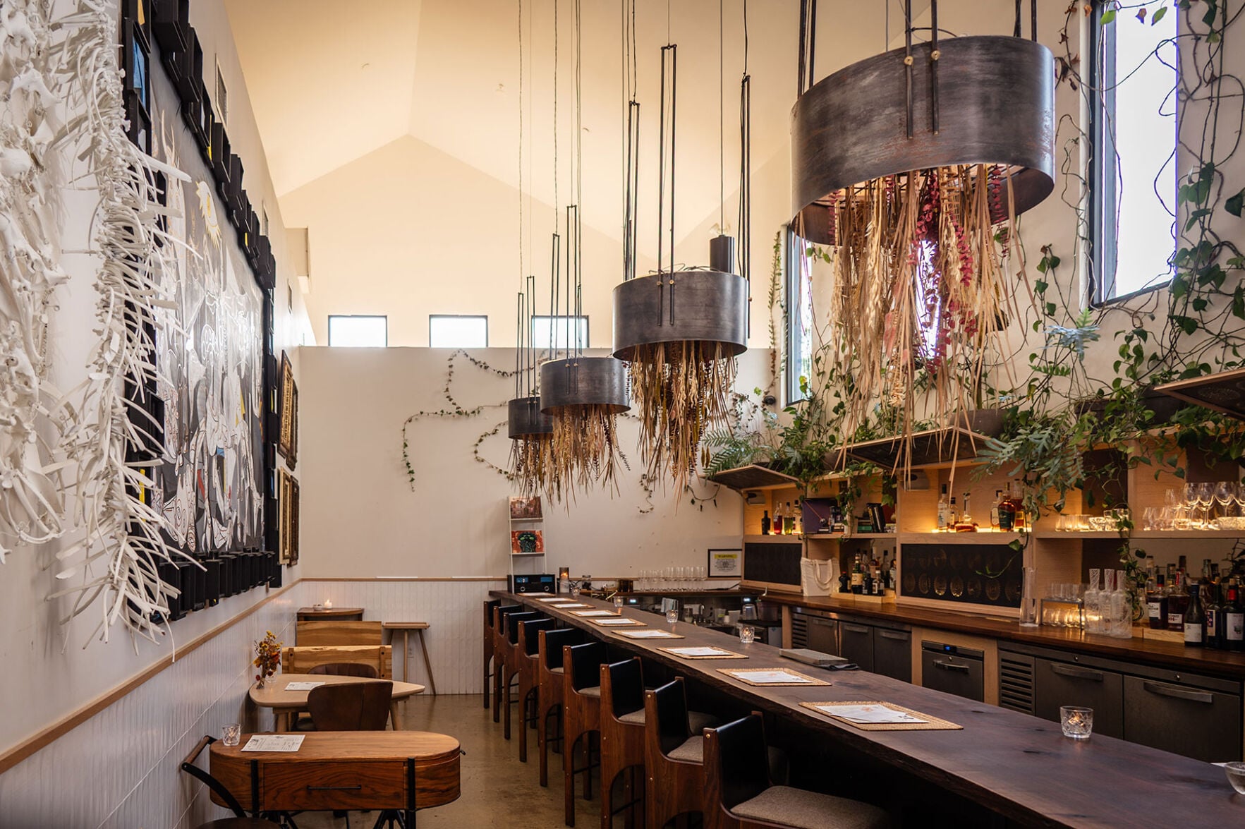 A line of stools in front of a bar with stalks of wheat hanging overhead from chandaliers.