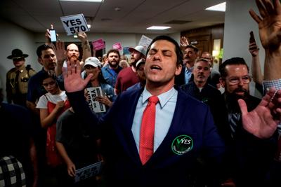 A man in a blue suit raises hands, a crowd behind him, one man waving a sign that reads “Yes 570”