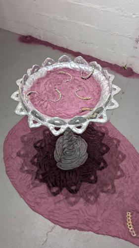 A silvery birdbath that’s been filled with rose-petal dust, surrounded by a beach of rose-petal dust that creates a teardrop shape on the floor around the sculpture.