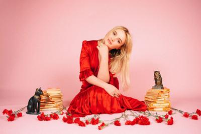 Emma Swift press photo 2025 artist in red dress sits flanked by stacks of books guarded by statues of an owl and a cat