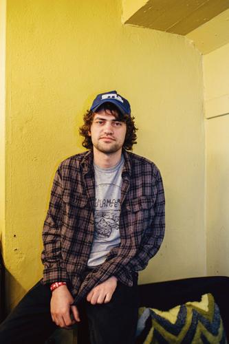 MJ Lenderman press photo artist sits on the arm of a striped couch in a room with yellow walls