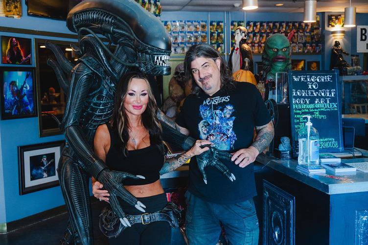 Stacey Dixon poses between the arms of a xenomorph from the Alien franchise next to Ben Dixon in a horror-themed tattoo parlor