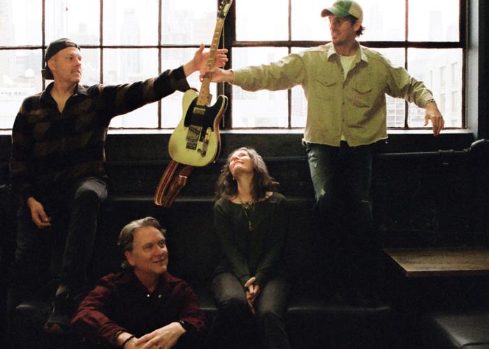Color press photo of the supergroup World Famous Pets, seated in front of a large window as one member passes another a Fender Telecaster guitar.