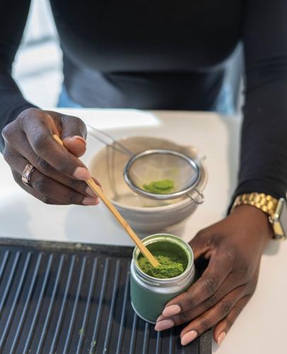 A woman's hands use a  thin wooden spoon to scoop green powder out of a jar and into a strainer that rets atop a ceramanic bowl