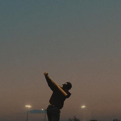 Photo of the artist on a playing field at sunset, facing away from the camera and swinging a bat as the park lights come on