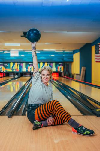 Color photo of Brennan Wedl sitting at the end of a bowling lane hoisting a bowling ball, in a pose resembling a bowling trophy