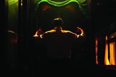A still from the film 'Resurrection' shows the back of a man standing arms raised above th shoulder, a green archway over his head leading into a dark hallway