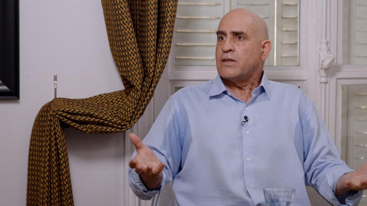 Daniel Siad speaking during an indoor interview, wearing a light blue button-up shirt and gesturing with both hands while seated