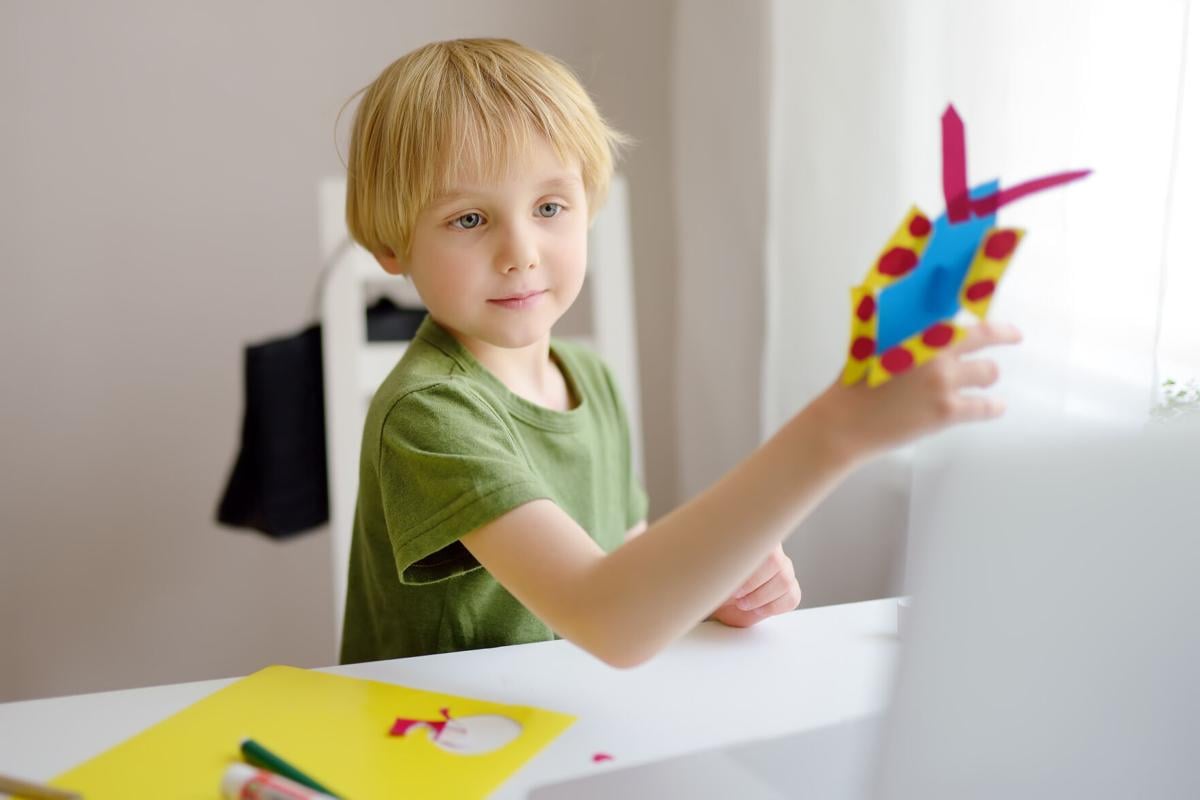 Boy making colored paper crafts at home during coronavirus lockdown. Child showing result teacher by laptop. Creativities lessons, distance learning, homeschooling, online kindergarten for kids.