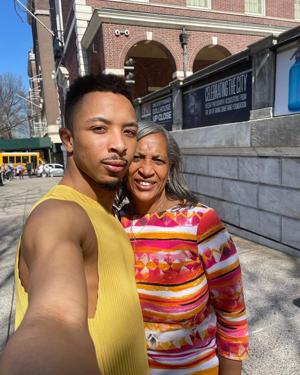 Image of Zelig Williams taking a selfie with his mother outdoors on a sidewalk. Williams is a light-skinned black man with a close-cropped black hair and mustache wearing a mustard-colored tank top.