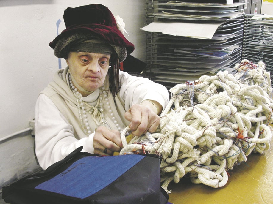 Judith Scott sits working on a fiber arts project. She wears multiple hats on her head and different pearl necklaces in varying sizes. She has a determined look on her face. 