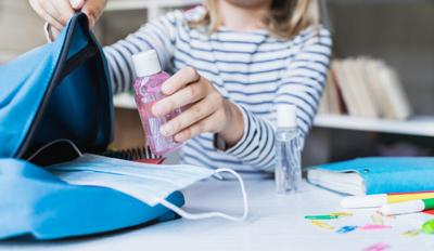 Little girl packing blue backpack in kids's room. Face mask, bottle of sanitizer, stationery, pens, multicolored markers, notebooks. Back to school. Mom's hygiene,safety precautions after coronavirus