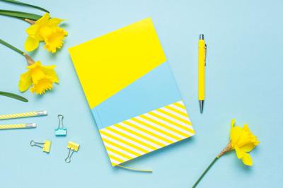 Yellow-blue notebook, pen, clips, spring flowers daffodils narcissus on blue background. Female desktop, Office desk, spring concept. Flat lay, top view, copy space. Template for feminine blog