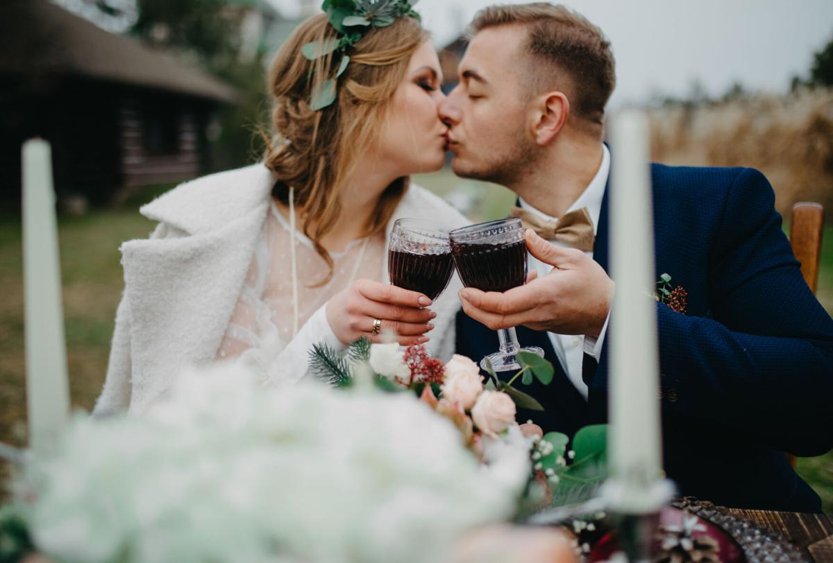 Newlyweds sit near banquet table, clink glasses of wine and kiss.