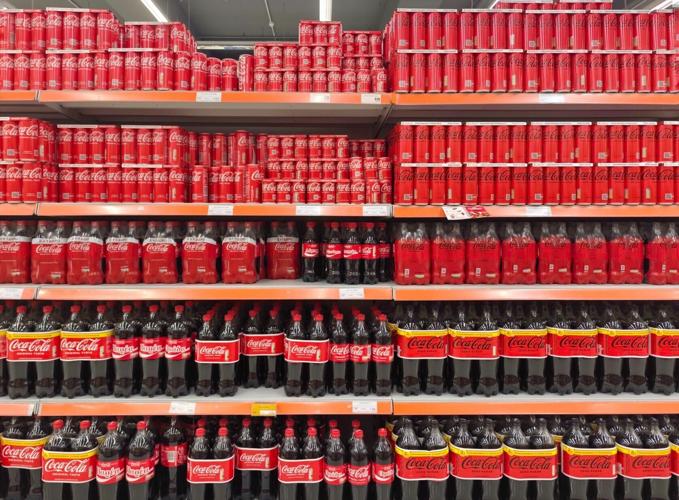 Photo of a fully stocked supermarket shelf filled with neatly arranged Coca-Cola cans and bottles in various sizes.