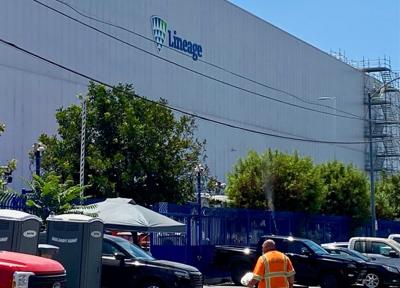Lineage warehouse behind blue fencing, parked vehicles and portable toilets, with a worker in an orange safety vest in the foreground.