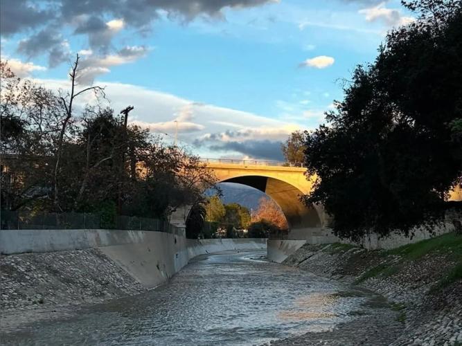Water flows through the Arroyo Seco under the York Boulevard Bridge in Hermon after heavy rains, with sunlight hitting the bridge’s arches and trees lining the concrete channel.