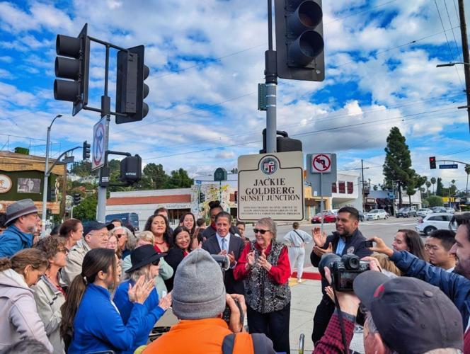 A group of people stand outdoors at a podium near a busy Silver Lake intersection, with newly unveiled street signs displayed behind them during a public ceremony.